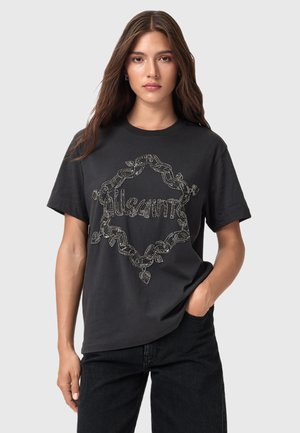 Mujer joven con cabello largo castaño, vistiendo una camiseta negra con un diseño de cadenas plateadas y hojas, y texto en la parte delantera, con una mano en el bolsillo.