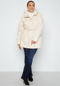 Světlobéžová voděodolná bunda s páskem na stažení v pase, zapínáním na zip a odnímatelnou kapucí. V kombinaci s tmavě modrými zvonovými džíny a černými podpatky.