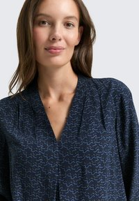 Navy blouse featuring a v-neck, soft texture, and a wavy blue abstract pattern. Loose fit with pleat detail at the shoulders.