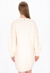 Cremefarvet strikket kjole i sweater-stil med ballonsleeves, strukturerede mønstre på ærmerne og ribkant. Har et lille mærke på bagsiden.
