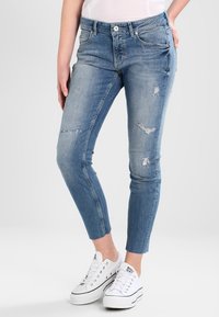 Vaqueros de denim azul ajustados con un ligero desgaste, lavado descolorido y una cintura de altura media. Combinados con zapatillas de lona blancas.
