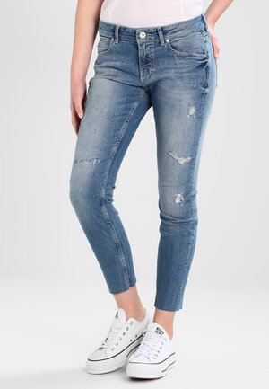Vaqueros de denim azul ajustados con un ligero desgaste, lavado descolorido y una cintura de altura media. Combinados con zapatillas de lona blancas.