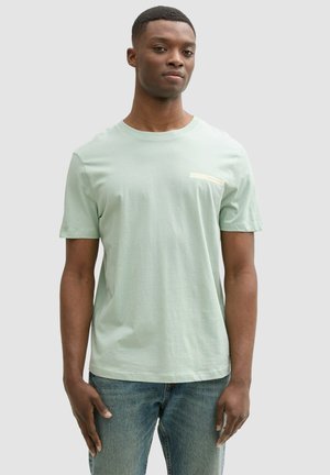 Camiseta de manga corta de color verde claro, fabricada con una tela suave, que presenta un pequeño bolsillo blanco en el lado izquierdo del pecho y un corte holgado.