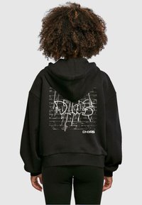 Svart oversized hoodie med en grafitti-inspirerad grafik på ryggen, med en texturerad tegelvägg som bakgrund och vit text.