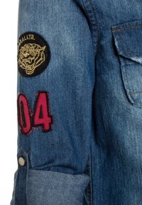 Camisa de denim de tejido azul, con parche de tigre negro y "04" bordado en rosa en la manga. Incluye detalle de puño abotonado.