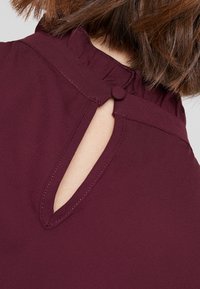 Top color bordeaux con scollo a volant, chiusura con bottoni e dettaglio a forma di chiave sulla schiena, realizzato in un tessuto liscio e leggero.