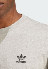 Siv pulover s krojenim ovratnikom, izdelan iz teksturiranega blaga. Ima majhen črn Adidas logo na levi strani in gladek ovratnik.