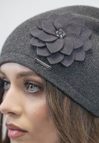 Berretto in maglia grigio con un grande fiore in feltro di un grigio più scuro, caratterizzato da petali sovrapposti e un centro in strass, abbinato a un'etichetta in metallo.