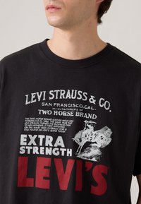 Vyras dėvi juodą Levi's marškinėlį su balta ir raudona tekstu bei grafika, kurioje pavaizduoti du žirgai, tempiantys odinį ženklelį.