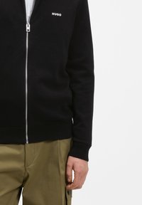 Άνδρας που φοράει μαύρο πουλόβερ με φερμουάρ και το λογότυπο "HUGO" στο στήθος και χακί παντελόνι cargo, στέκεται μπροστά σε λευκό φόντο.