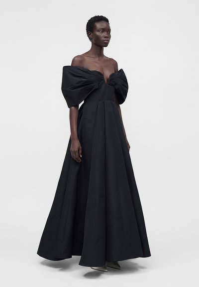 Γυναίκα που φοράει μαύρο βραδινό φόρεμα με ακάλυπτους ώμους, υπερμεγέθη φουσκωτά μανίκια και μακριά φούστα, στέκεται μπροστά από απλό φόντο.