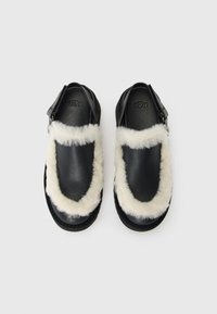 Zwarte instappers met een witte, pluizige bontafwerking die de voorkant en enkels van de bandjes omringt, met het UGG-logo op de binnenzool van elke sandaal.