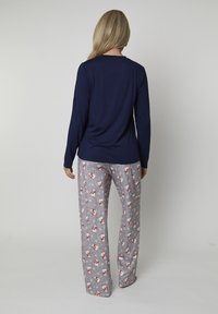 Donna con lunghi capelli biondi che indossa un maglione navy a maniche lunghe e pantaloni da pigiama grigi con motivi di pupazzi di neve rossi e bianchi, in piedi di spalle.