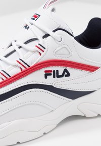 Zapatilla blanca con upper de cuero texturizado, acentos en rojo y navy, suela acolchada y logo "FILA" bordado. Incluye cordones ajustables.