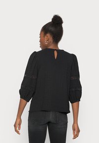 Blusa preta com mangas bufantes, detalhes em renda nas mangas e um decote em forma de gota nas costas. O tecido possui um padrão texturizado e é leve.