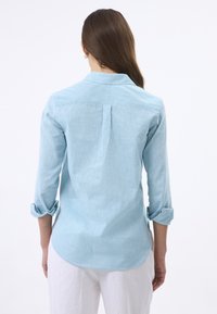 Camisa de manga larga de color azul claro, con cuello, puños enrollados y una plisada en la parte trasera. Textura suave, dobladillo curvado y corte relajado.