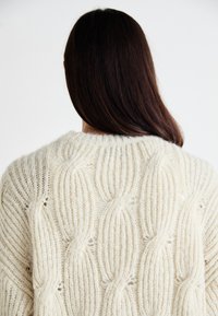 Weißer strukturierter Pullover mit einem Zopfmuster und rundem Halsausschnitt, der offene Strickdetails entlang des Rückens zeigt.