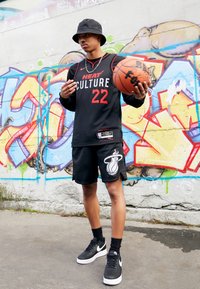 Fekete kosárlabdámez, rajta "HEAT CULTURE 22" felirattal piros színben, fekete rövidnadrág fehér logóval, fekete kalap, kosárlabdát tartva, városi graffiti háttér.