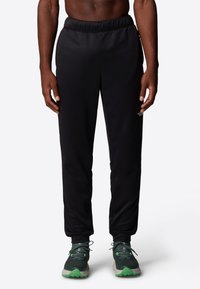 Black athletic pants with an elastic waistband, featuring a logo on the left side, paired with green-accented running shoes.
