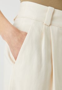 Világos bézs nadrág sima, texturált anyagból, melynek elején plisszált rész és oldalzsebek találhatók, gondosan szabott, testhez álló derekú.