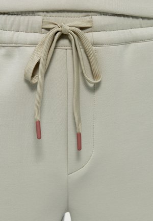 Nahaufnahme des Taillenbunds einer beigefarbenen Jogginghose mit gebundenem Kordelzug, der pinke Kunststoffspitzen hat, und sichtbarer Seitentaschennähte.