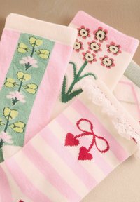 Tres pares de calcetines de colores pasteles con diseños de flores y corazones, incluyendo detalles de encaje y rayas rosas, colocados sobre una superficie beige.