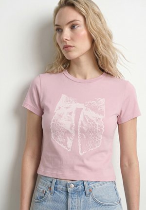 Blondynka ubrana w jasnoróżowy krótki t-shirt z grafiką białej koronkowej kokardki, w połączeniu z jasnoniebieskimi jeansami z wysokim stanem na jednolitym tle.