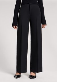 Kobieta ubrana w czarne, szerokie spodnie o wysokim stanie z frontowymi kantami oraz czarne, lakierowane buty na obcasie w kształcie migdała.