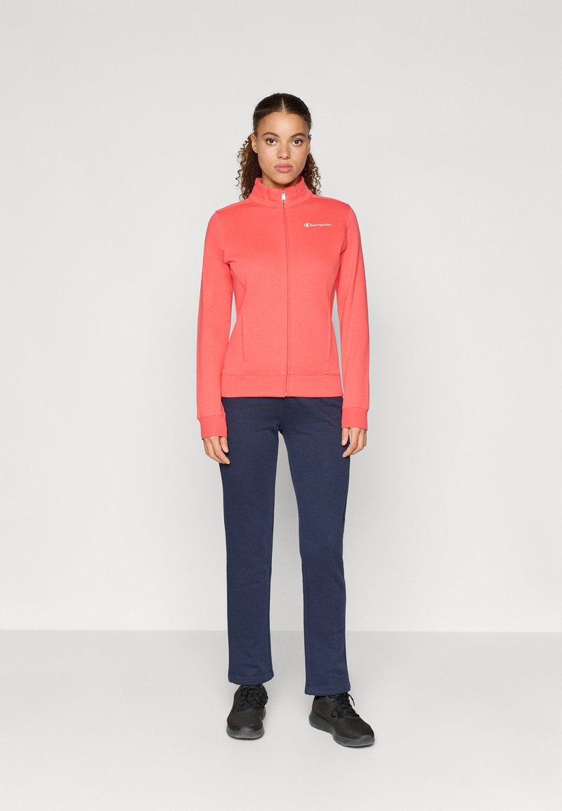 Hanorac cremos cu fermoar, cu guler înalt și logo, asociat cu pantaloni bleumarin și adidași negri. Materialul pare moale, cu un design ajustat.