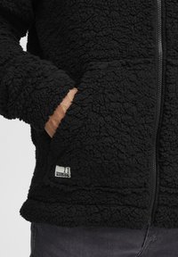 Hand in pocket of black textured fleece jacket with front zipper and small white "11 Project" label near pocket.