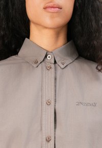Gray shirt with a button-down collar, brown buttons, and embroidered "2NDDAY" logo. Smooth texture, tailored fit, and subtle shine.