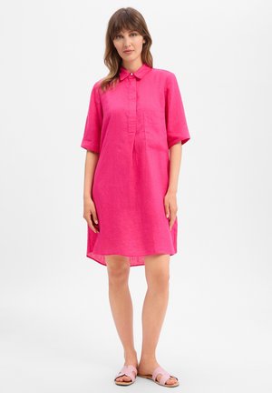 Femme portant une robe chemise manches courtes rose vif avec col et poche avant, associée à des sandales slides rose clair, debout sur fond blanc.