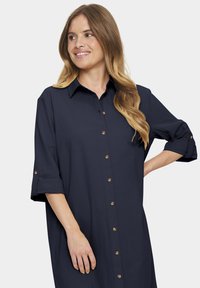 Rochie bleumarin cu guler, închidere frontală cu nasturi și mâneci răsucite. Prezintă nasturi din lemn și o croială lejeră. Textura materialului este moale.