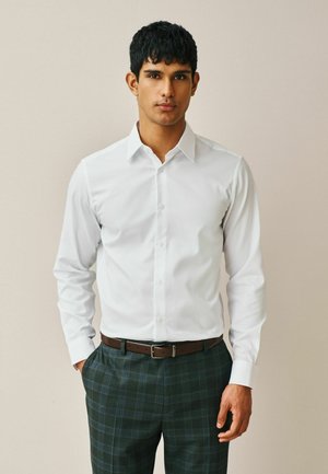 Junger Mann mit kurzen dunklen Haaren, trägt ein weißes Hemd mit Knöpfen, das in grüne Karohosen mit einem braunen Ledergürtel gesteckt ist.