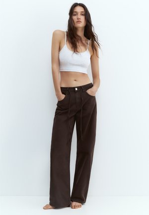 Fiatal nő hosszú sötét hajjal, fehér crop topot és laza sötétbarna nadrágot visel, mezítláb áll fehér háttér előtt.