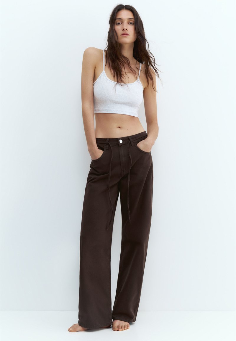 Junge Frau mit langen dunklen Haaren trägt ein weißes Crop-Top und lockere dunkelbraune Hosen, steht barfuß vor weißem Hintergrund.