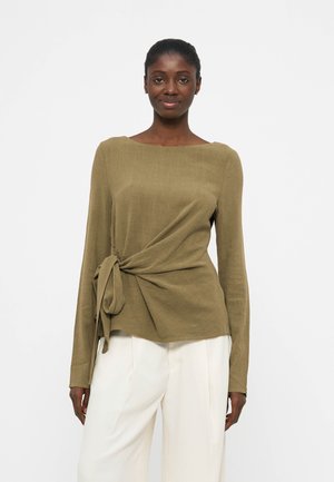 Γυναίκα που φοράει ελιάς μακρυμάνικη μπλούζα με λεπτομέρεια δεσίματος στο πλάι και λευκό παντελόνι φαρδιάς μίνης, στέκεται μπροστά σε έναν απλό λευκό φόντο.