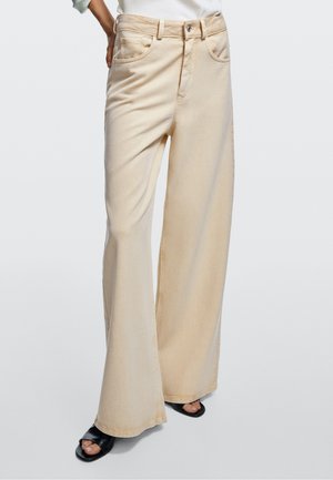 Pantalon beige à jambes larges porté avec des chaussures noires à bouts ouverts, montré de la taille aux pieds sur un fond uni.