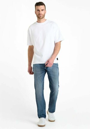 T-shirt en coton blanc avec une coupe décontractée, associée à un jean en denim à lavage moyen. La tenue est complétée par des baskets blanches avec une semelle beige.