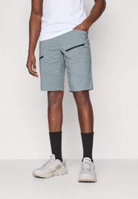 Shorts gris clair avec deux poches zippées à l'avant, fabriqués dans un tissu léger. Portés avec un haut blanc et des baskets grises.