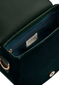 Mała, ciemnozielona torebka z weluru z złotym zapięciem i wewnętrzną kieszonką na zamek, oznaczona napisem "Fable England", pokazana od góry z otwartym wnętrzem.