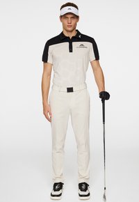 Golfpolo i ljusgrått med svarta accenter, med ett hexagonmönster; beige byxor och svart golfhandske; person som håller en golfklubba.