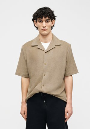 Νέος άνδρας με σκούρα σγουρά μαλλιά φοράει μια μπεζ κοντομάνικη πουκάμισα με κουμπιά πάνω από ένα λευκό εσώρουχο και μαύρο παντελόνι, στέκεται μπροστά σε ένα απλό φόντο.