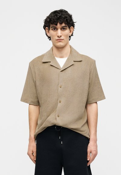 Joven con cabello rizado oscuro lleva una camisa beige de manga corta abotonada sobre una camiseta blanca y pantalones negros, posando contra un fondo liso.