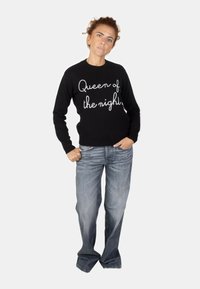 Maglione nero lavorato a maglia con "Regina della notte" in bianco corsivo, abbinato a jeans in denim azzurro chiaro, in piedi contro uno sfondo grigio uniforme.
