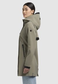 Cappotto impermeabile color khaki con cappuccio, tasche con zip e dettagli neri. Texture liscia, taglio dritto e caratteristiche regolabili.