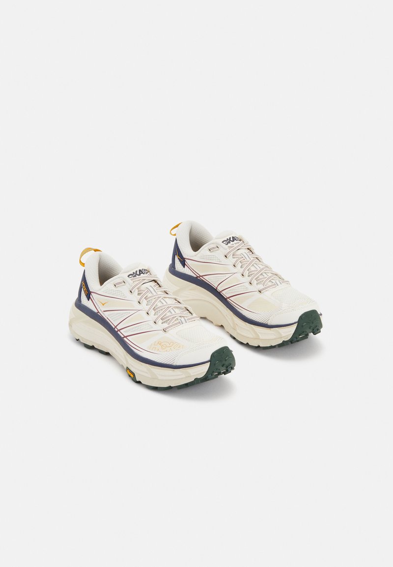 White running shoes with mesh upper, contrasting navy accents, purple detailing, and a rugged green outsole. Features stitched lines and a pull tab.