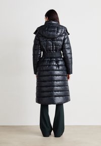 Μαύρο, επενδεδυμένο μακρύ παλτό με ζώνη στη μέση και κουκούλα, χαρακτηρισμένο από γυαλιστερή επιφάνεια και σχεδίαση με ραφές. Φορεμένο πάνω από σκούρους πράσινους παντελόνες.