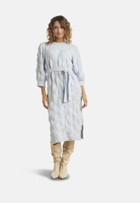 Frau posiert in einem hellblauen, strukturierten Strickkleid mit Gürtel, kombiniert mit beige-farbigen, lässigen kniehohen Stiefeln, vor einem schlichten weißen Hintergrund.