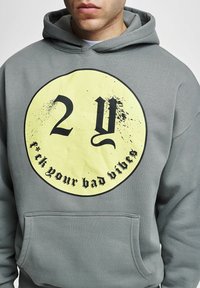 Lys grå hettegenser med en stor gul sirkelgrafikk med svart tekst som leser "2 Y" og "f**k dine dårlige vibber." Hette-design, frontlomme.